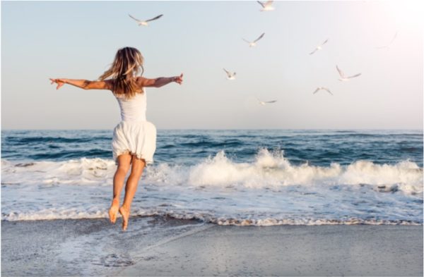A young girl jumps on the beach while a small wave crashes on shore.