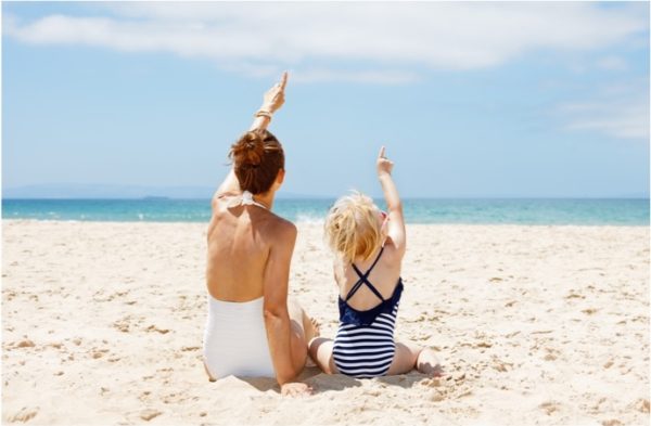 A mother and young child, sitting on the beach pointing to clouds in the sky.