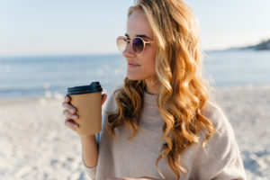 Romantic girl with long blonde hair drinking tea at sea. Outdoor photo of charming woman in sunglasses looking at ocean in autumn morning
