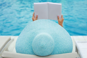 Beautiful young woman reading book while relaxing on sun lounger near swimming pool