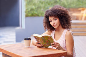 Young mixed race woman sitting a table outdoors with her takeaway coffee reading a book
