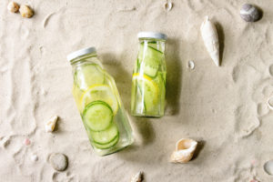 Summer drinks theme. Two glass bottles with lemon and cucumber infusion sassy water on white sand as background. Flat lay, space