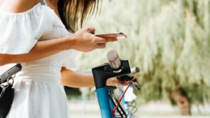 Side view of young woman paying rent of electric scooter by mobile app, outdoors. Close-up of girl in white dress using smartphone to pay for vehicle. Selective focus on female hand and phone.
