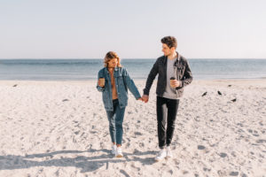 Two people holding hands and coffee while walking on the beach in the morning