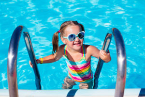 Happy laughing little girl playing in outdoor swimming pool on a hot summer day. Kid in colorful bathing suit and goggles learning to swim in tropical resort. Water fun for children