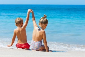 Happy kids have fun in sea surf on white sand beach. Couple of children sit in water pool with hands up. Travel lifestyle, swimming activities in family summer camp. Vacations on tropical island.