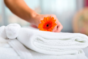 Maid doing room service in hotel, she is making up the beds. Stack of white towels with orange flower.