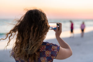 Young millennial girl woman taking landscape photo with phone smartphone of people in background at colorful sunset at Siesta Key Beach in Sarasota, Florida