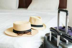 Luggage and beach hat of a couple on bed in modern hotel room with windows, curtains. Travel, relaxation, journey, trip and vacation concepts. Closeup