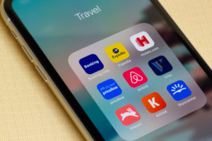 Assorted travel apps are seen on an iPhone on a lounge chair - Booking.com, Expedia, Hotels.com, Priceline, Airbnb, Vrbo, Hopper, KAYAK, and Skyscanner.